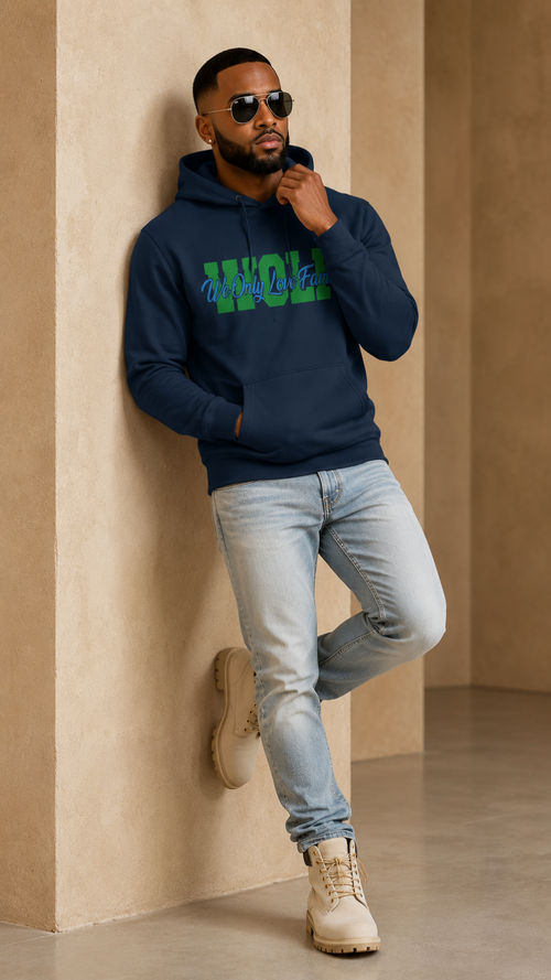 Man wearing a WOLF navy hoodie with green text, light blue jeans, and beige boots leaning against a beige wall.